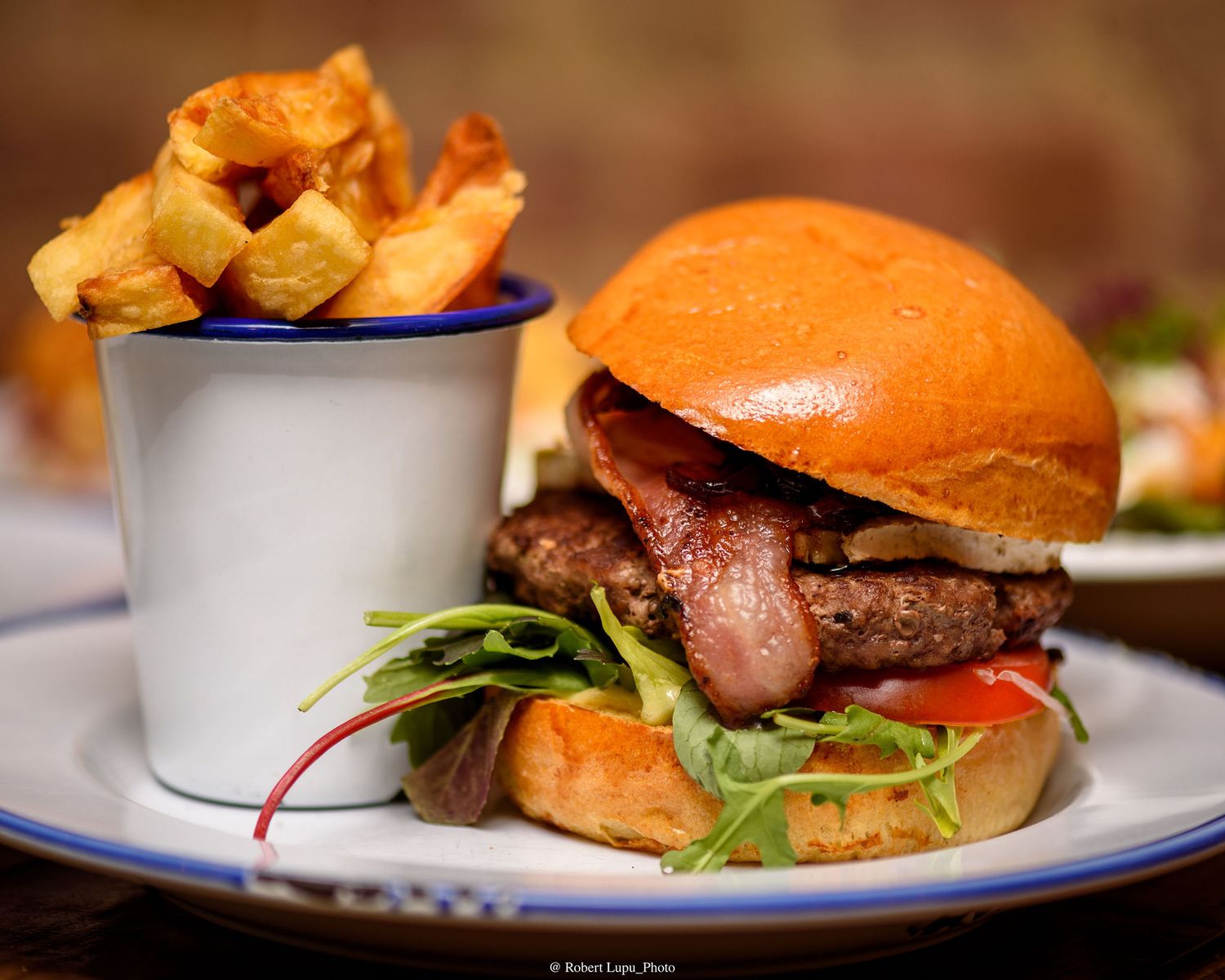 Gourmet burger with bacon, fresh greens, and tomato on a brioche bun, served with chunky fries in a metal cup.