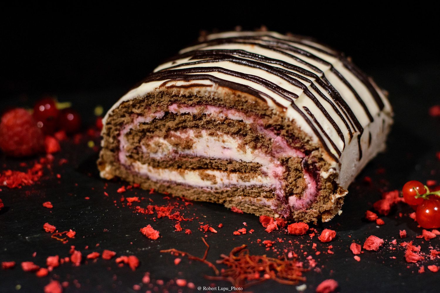 Chocolate raspberry Swiss roll cake with white cream filling, drizzled with dark chocolate stripes on a dark background.