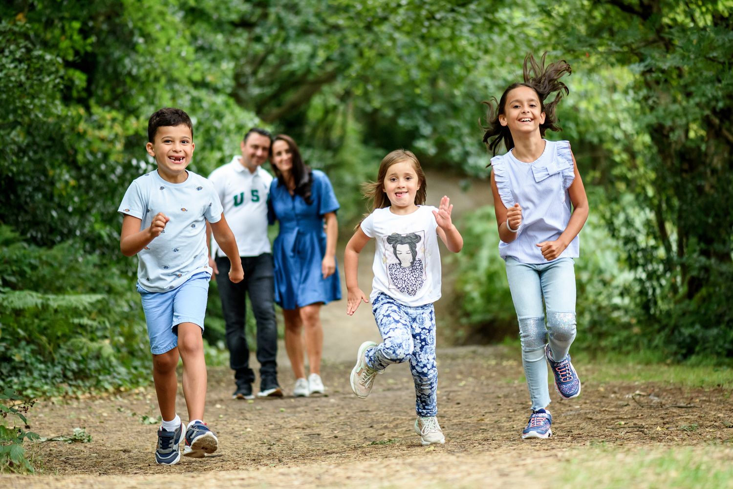 Family with two adults and three children running and playing on a sunny forest trail surrounded by green trees.