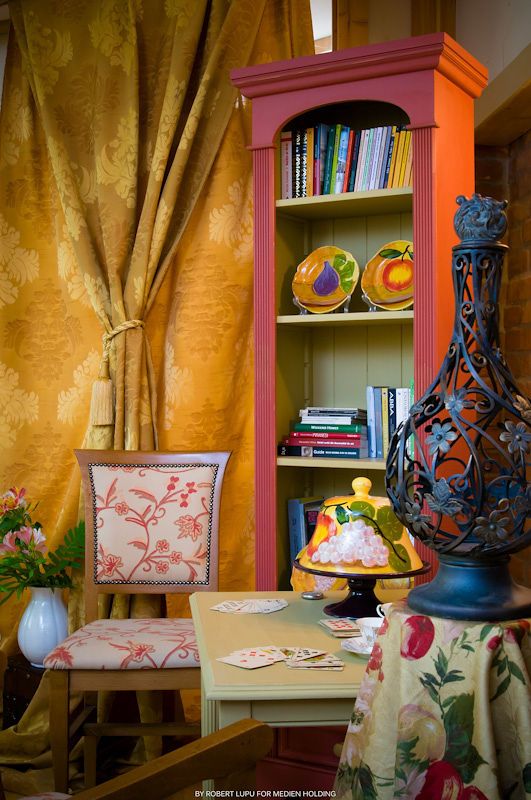 Colorful bohemian interior featuring a red bookcase, gold damask curtains, floral chair, and ornate black vase on a table.