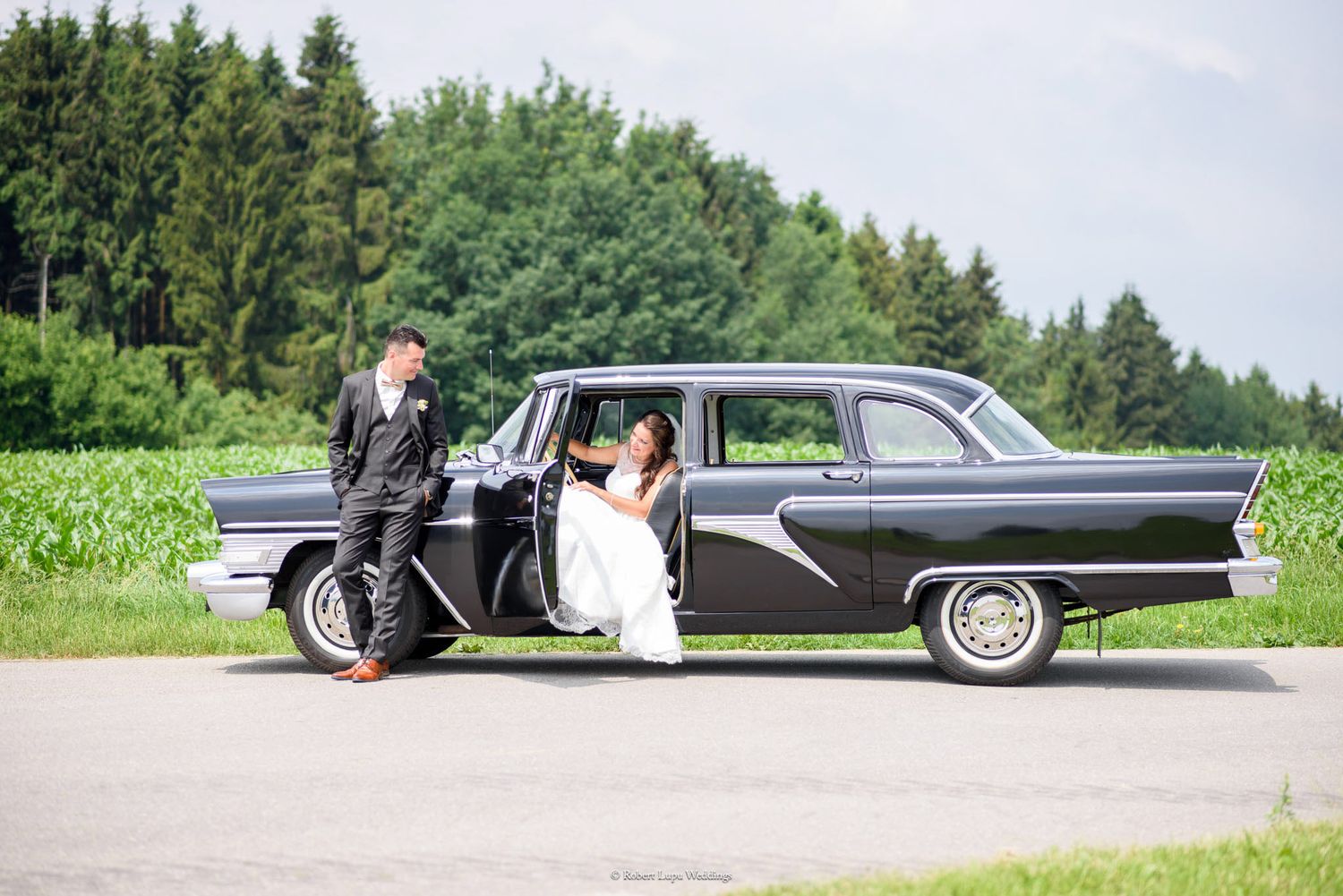 Bride and groom posing with a vintage black classic car on their wedding day.