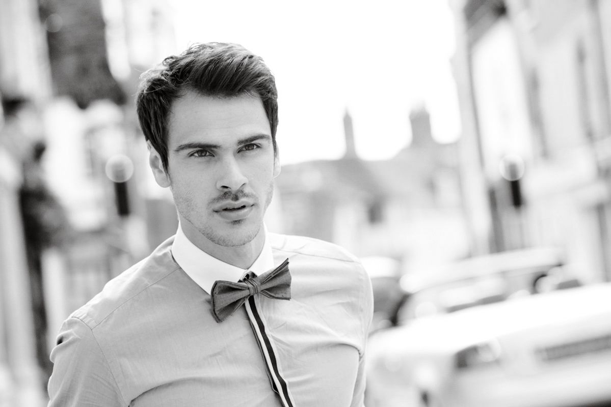 Stylish young man in white dress shirt with bow tie poses outdoors in black and white fashion photo.