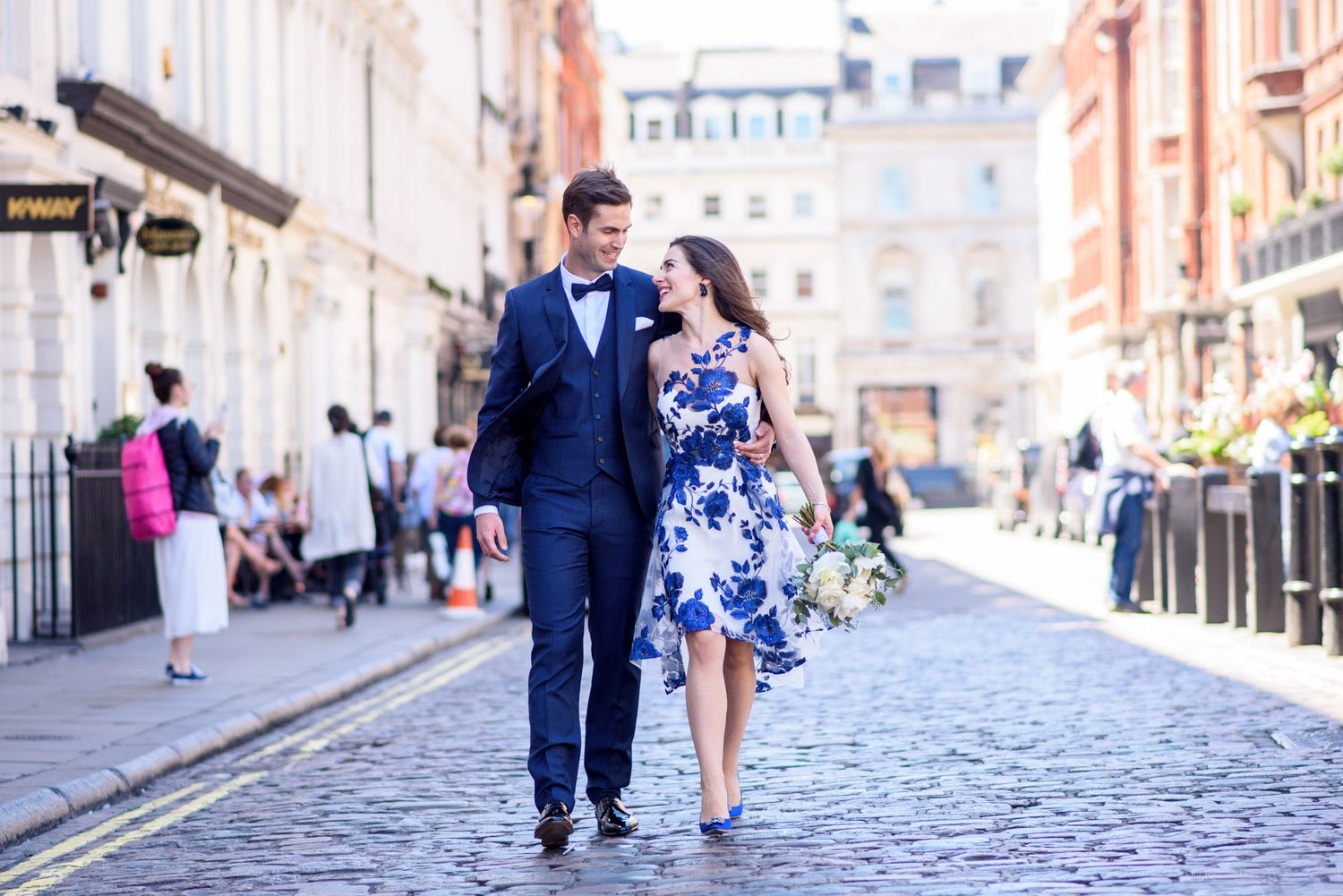 Couple in formal attire walking on cobblestone street in London, woman in blue floral dress, man in navy suit.