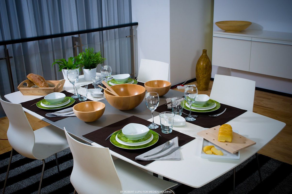 Elegant dining table set for six with green plates, wooden bowls, wine glasses, and white modern chairs in a stylish interior.