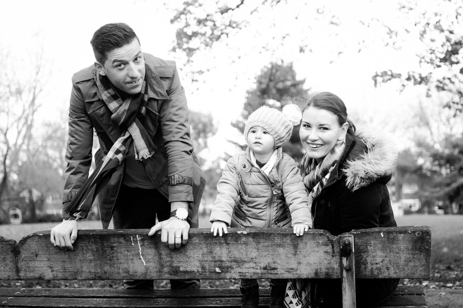 Black and white family portrait of parents and toddler leaning on wooden fence in autumn park.