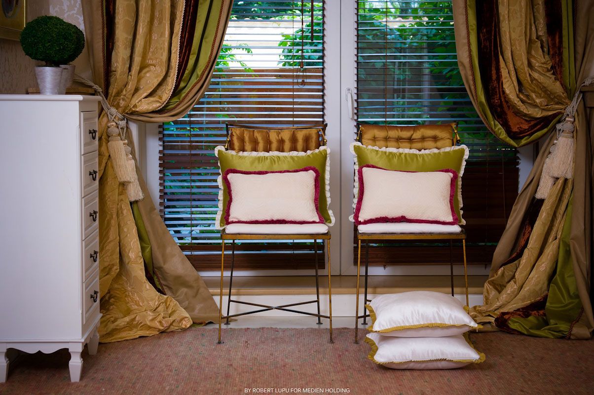 Two elegant chairs with decorative pillows sit between gold silk curtains near a window with wooden blinds.