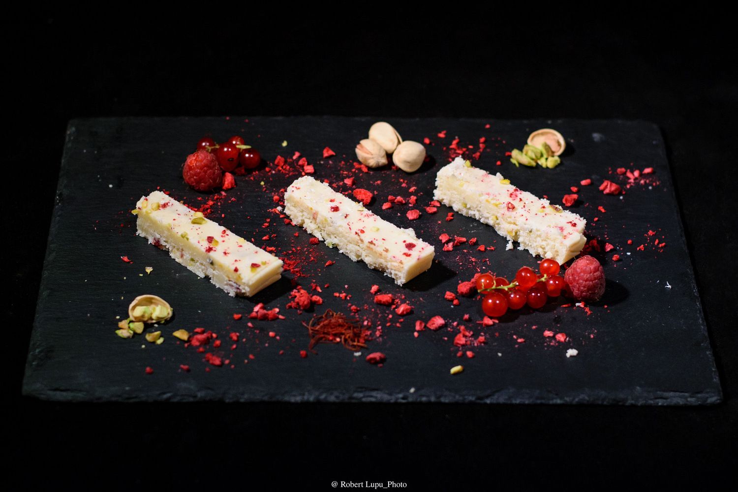Three rectangular white chocolate dessert bars garnished with freeze-dried raspberries and pistachios on a dark slate.
