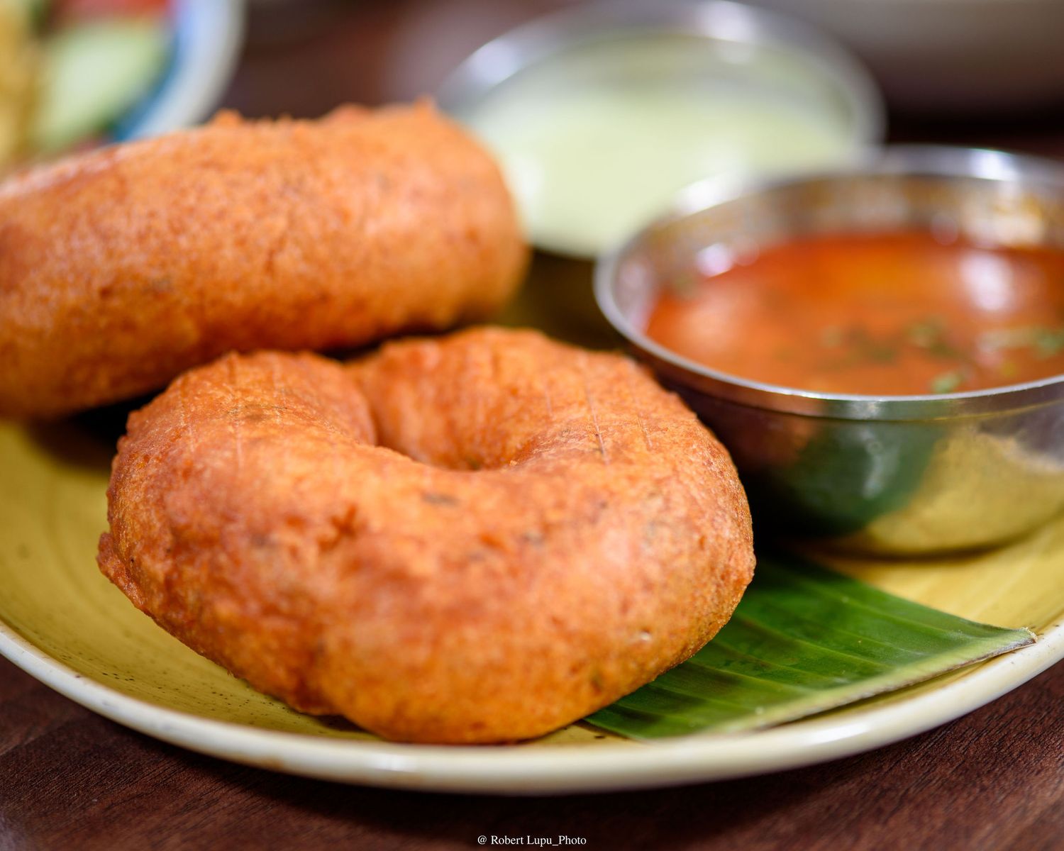 Two golden crispy medu vada donuts served on banana leaf with sambar and coconut chutney dipping sauces.
