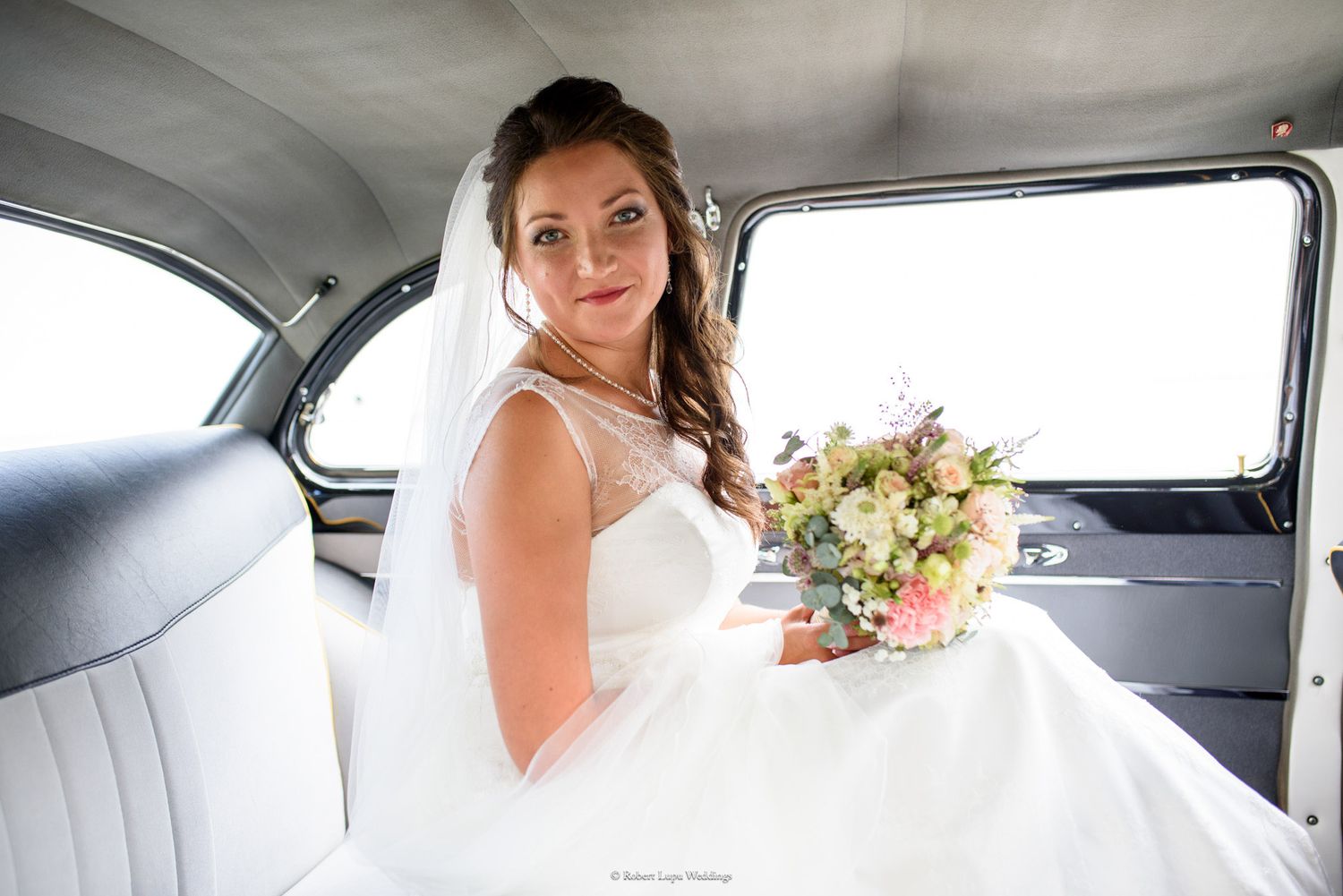 Bride in white gown holding a pastel bouquet seated in a vintage car on her wedding day.
