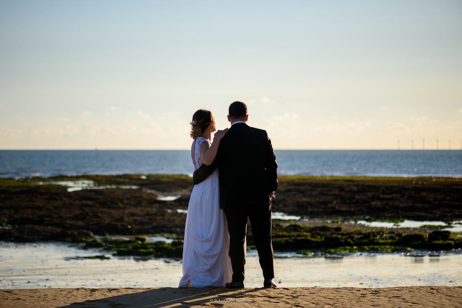 Bride and groom stand together on a sandy beach at sunset, gazing at the calm ocean horizon.