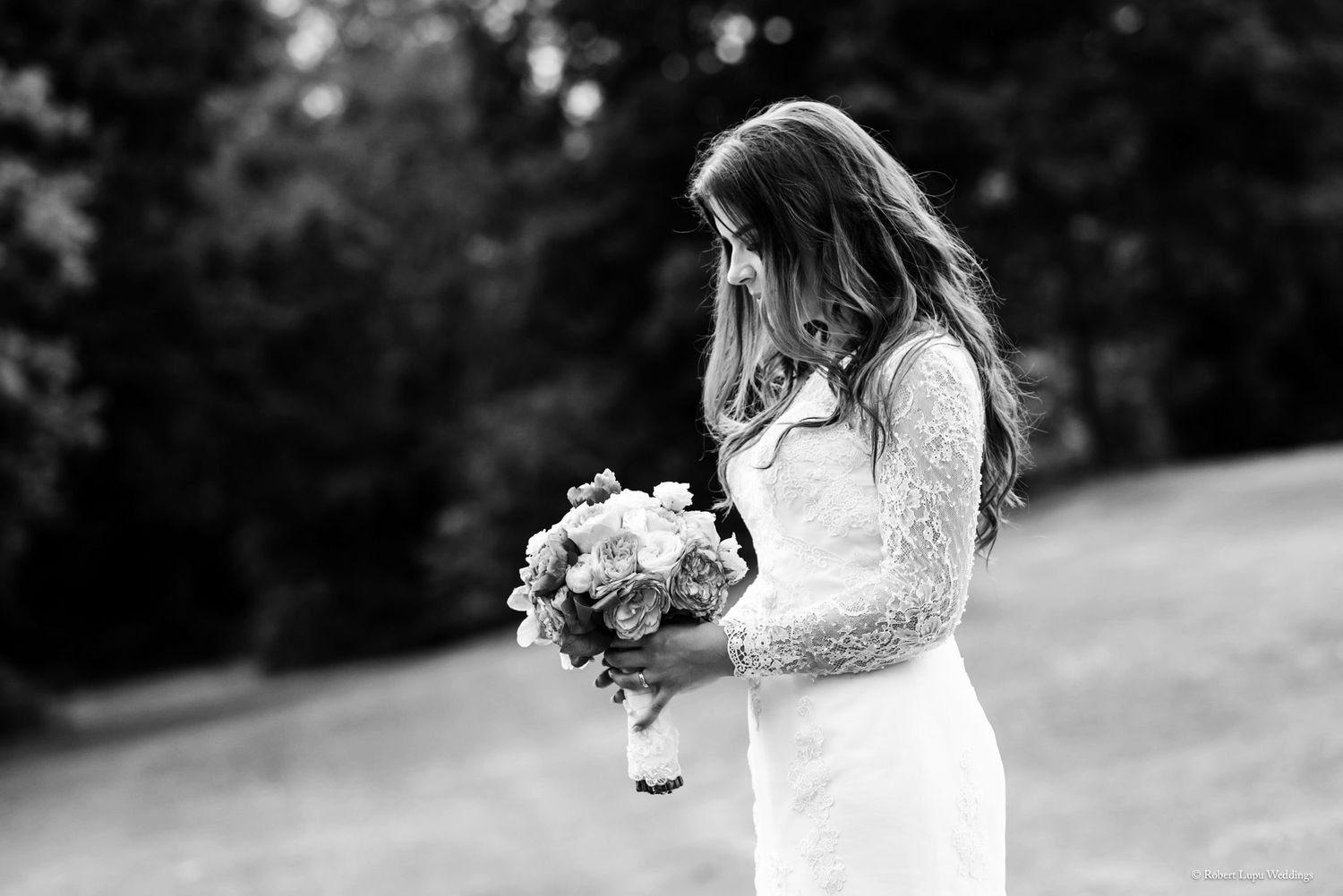 Black and white photo of a bride in a lace gown holding a bouquet, hair blowing in the wind outdoors.