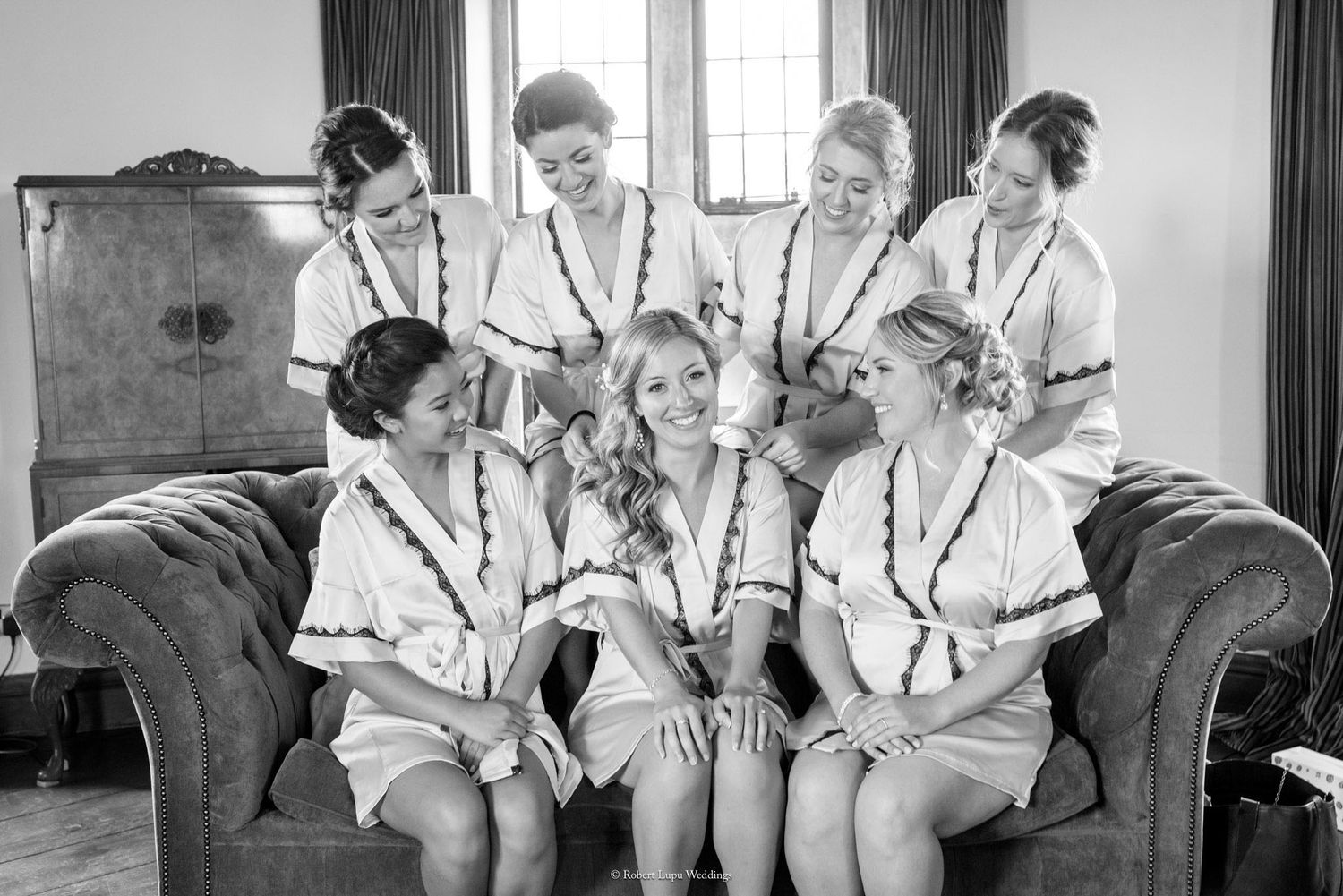 Bridesmaids in matching white robes laughing together on a vintage sofa in a black and white wedding preparation photo.