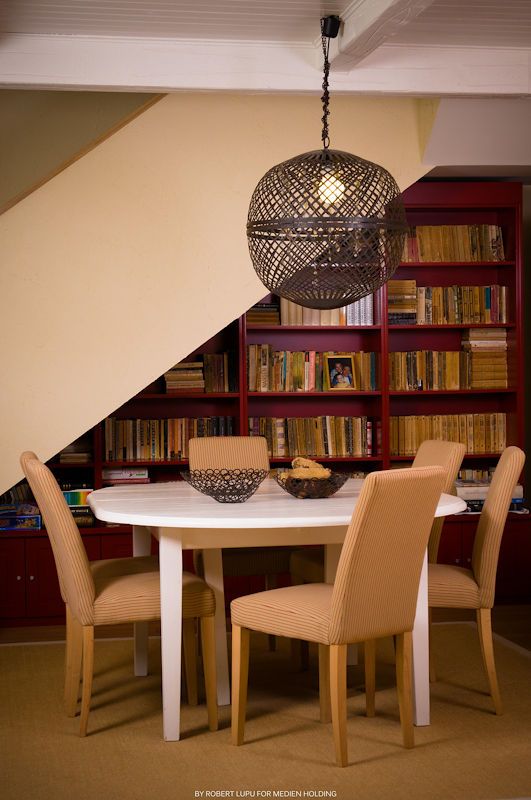 Round white dining table with tan chairs under ornate pendant light, beside red bookshelf filled with books.