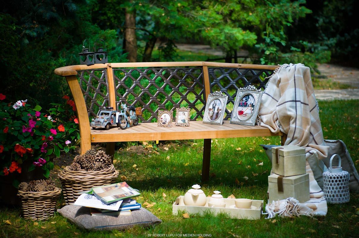 Wooden garden bench decorated with backpack, toy car, mugs, and blanket, surrounded by wicker baskets and flowers.