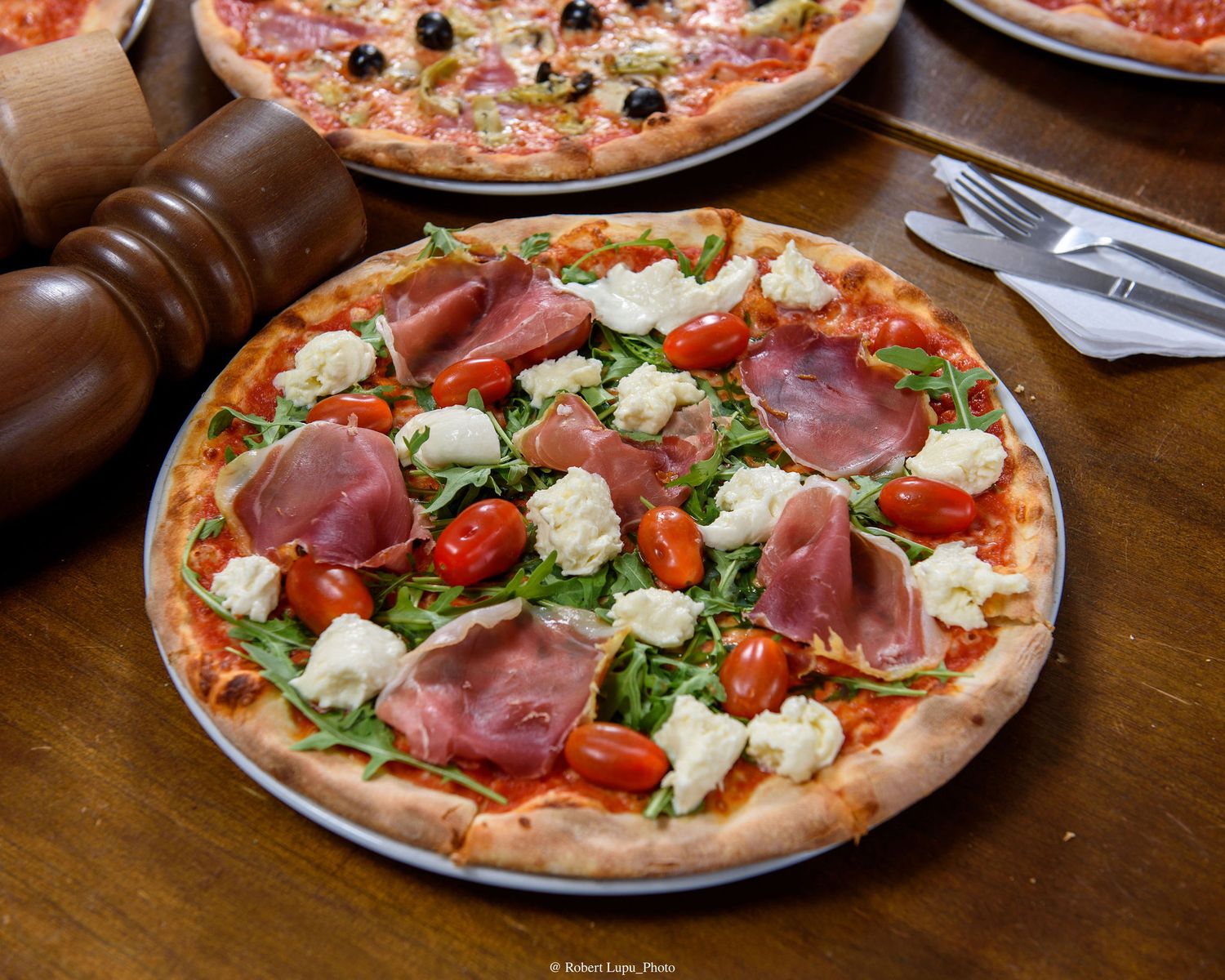 Italian pizza with prosciutto, cherry tomatoes, fresh mozzarella, and arugula on a wooden restaurant table.
