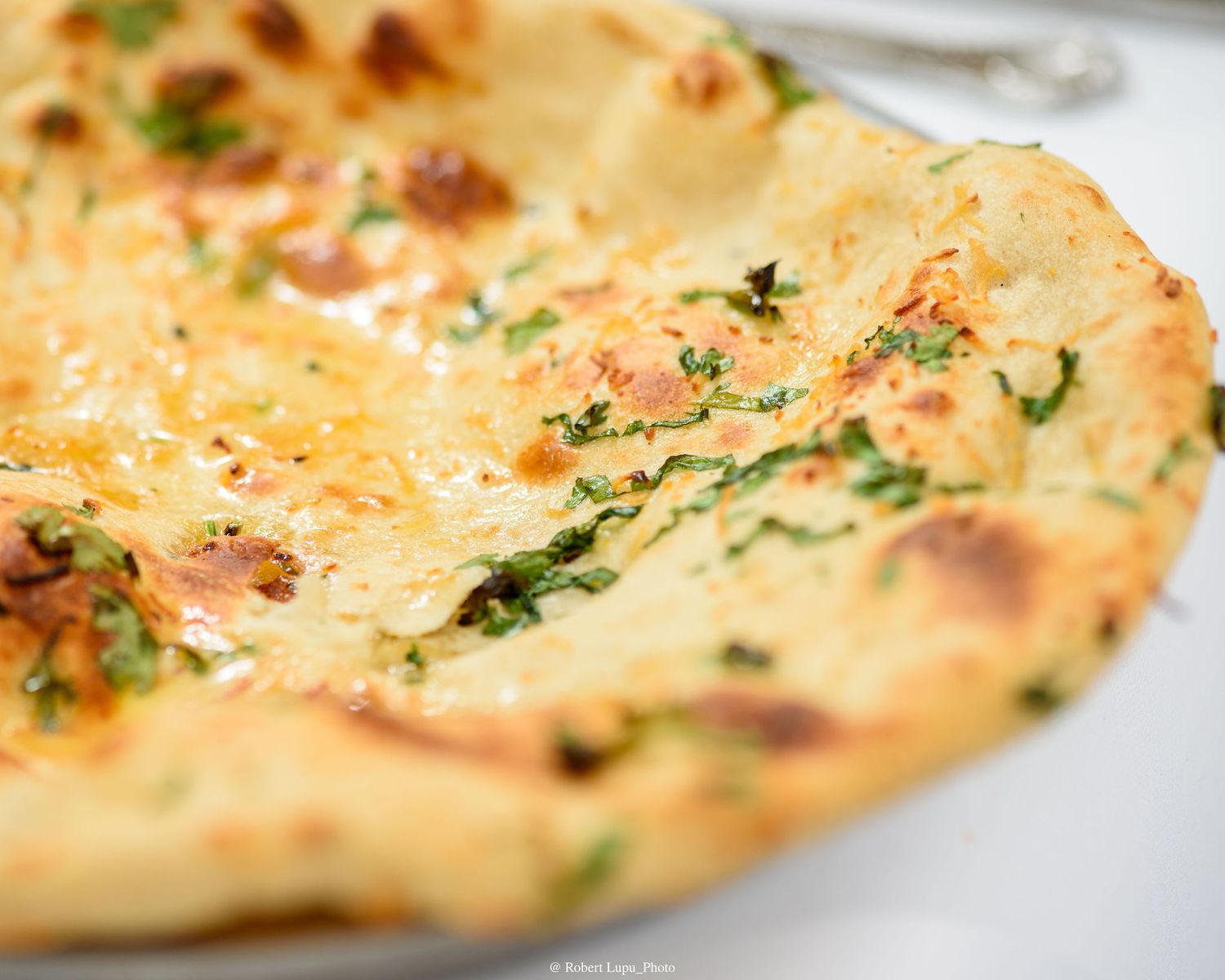 Close-up of a golden flatbread topped with melted cheese, fresh herbs, and bacon bits on a white plate.