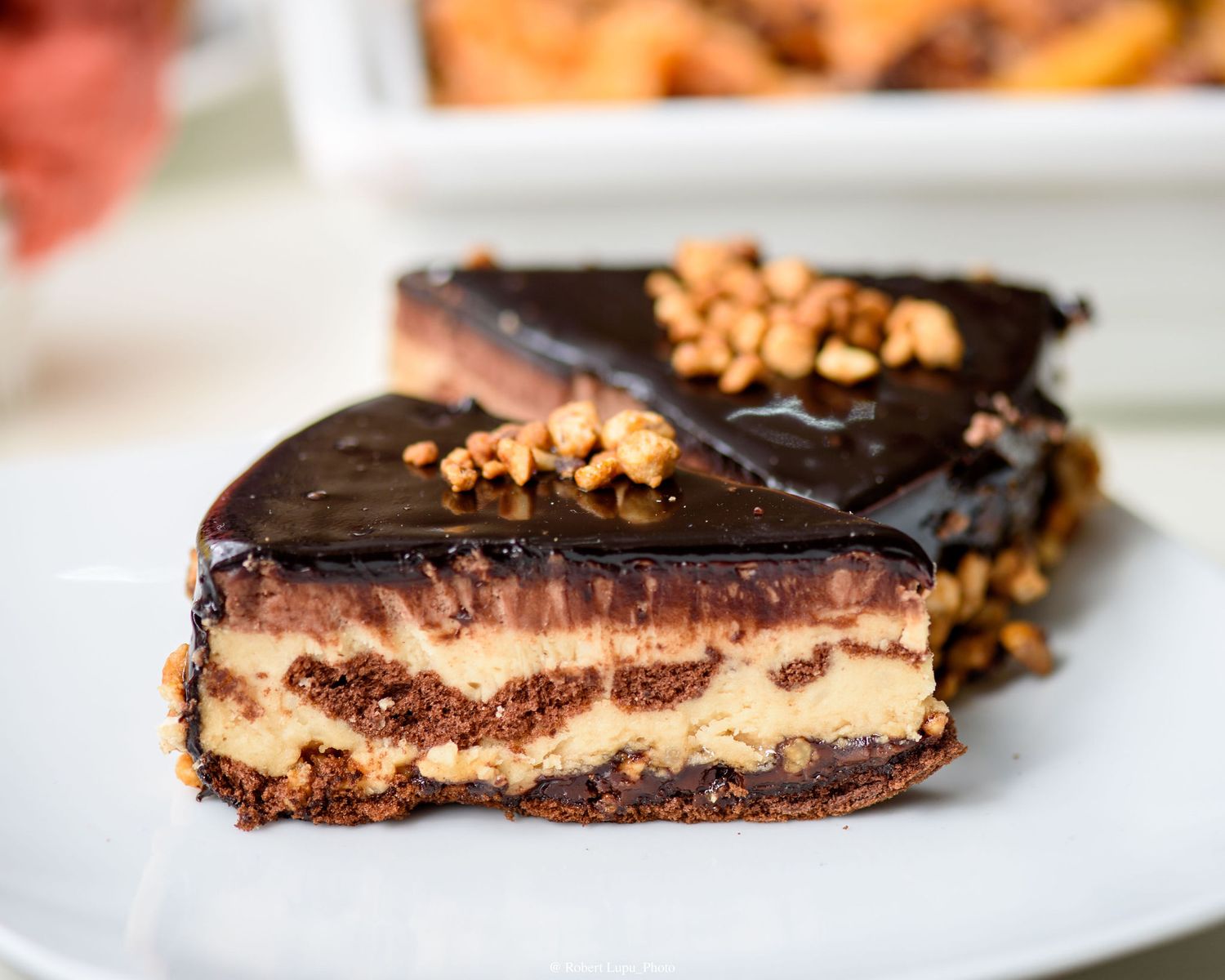 Decadent chocolate ganache layered dessert bar with peanut butter filling and chopped nuts on a white plate.