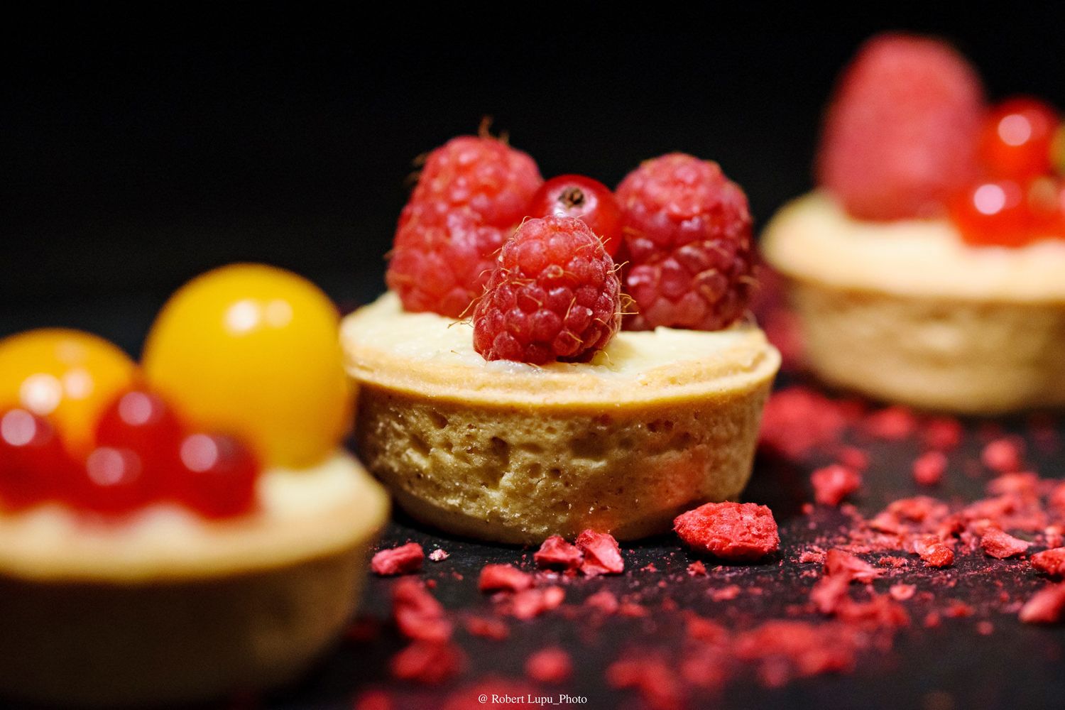 Close-up of mini fruit tarts topped with fresh raspberries and red currants on a dark background.