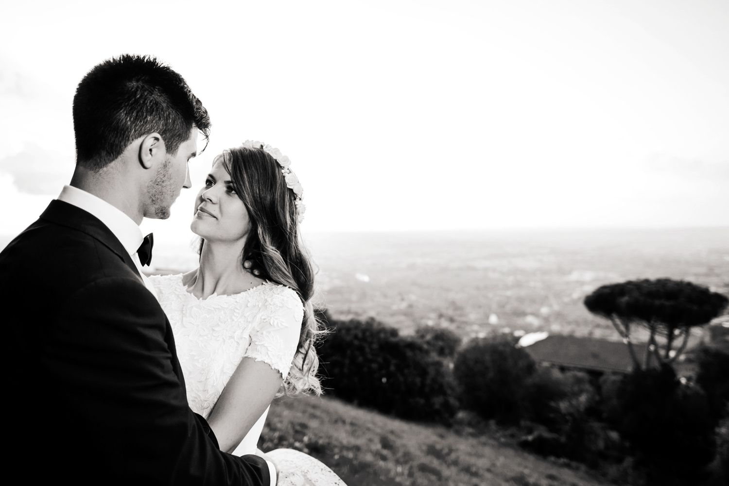 Black and white photo of a bride and groom embracing with a scenic cityscape and trees in the background.