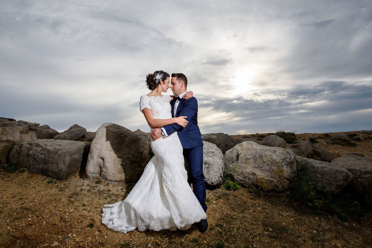 Bride and groom share a romantic kiss outdoors among large rocks under a dramatic cloudy sky on their wedding day.