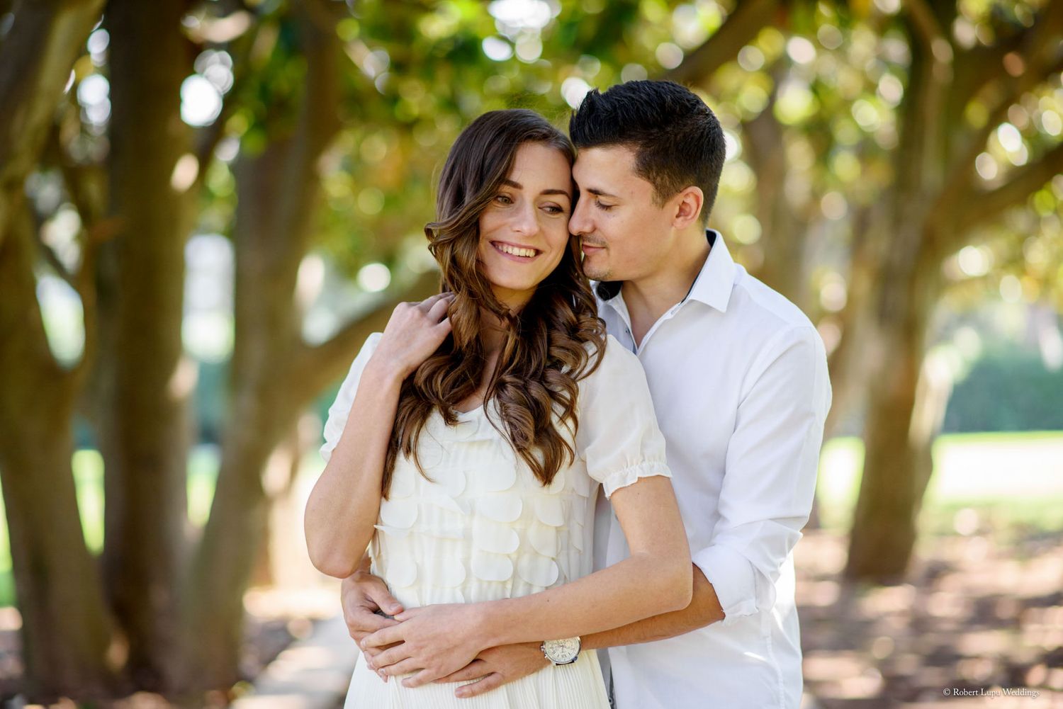 A couple embraces outdoors under trees, woman in white dress smiling as man holds her close in a romantic engagement photo.