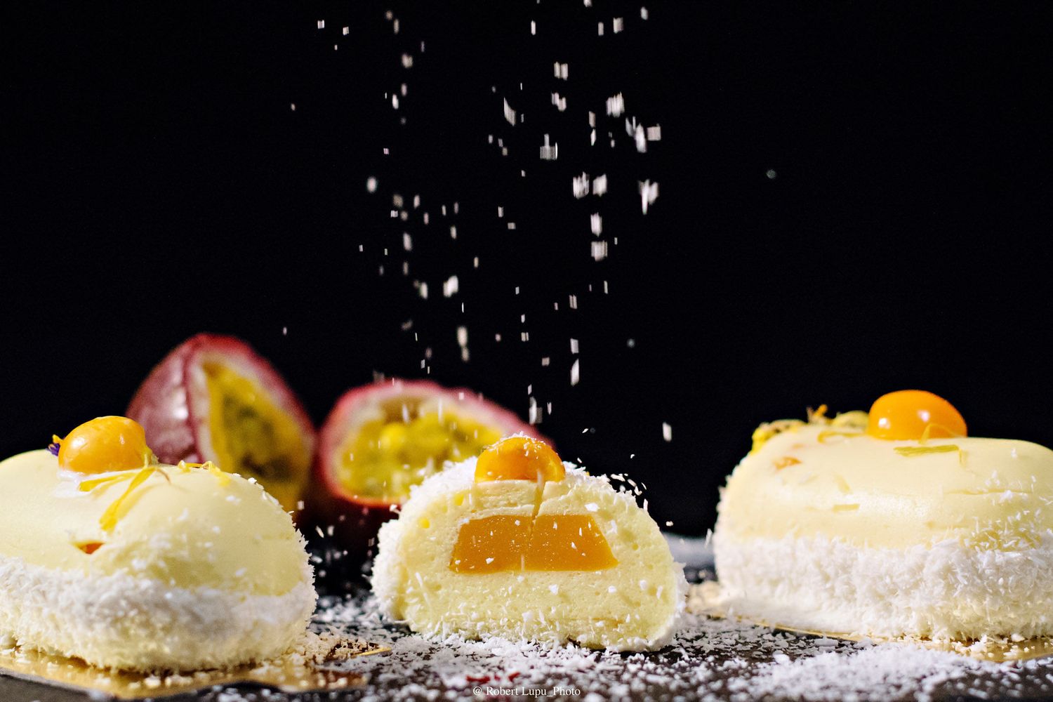 Coconut-dusted passion fruit mousse cakes with golden kumquats, powdered sugar falling dramatically against black background.