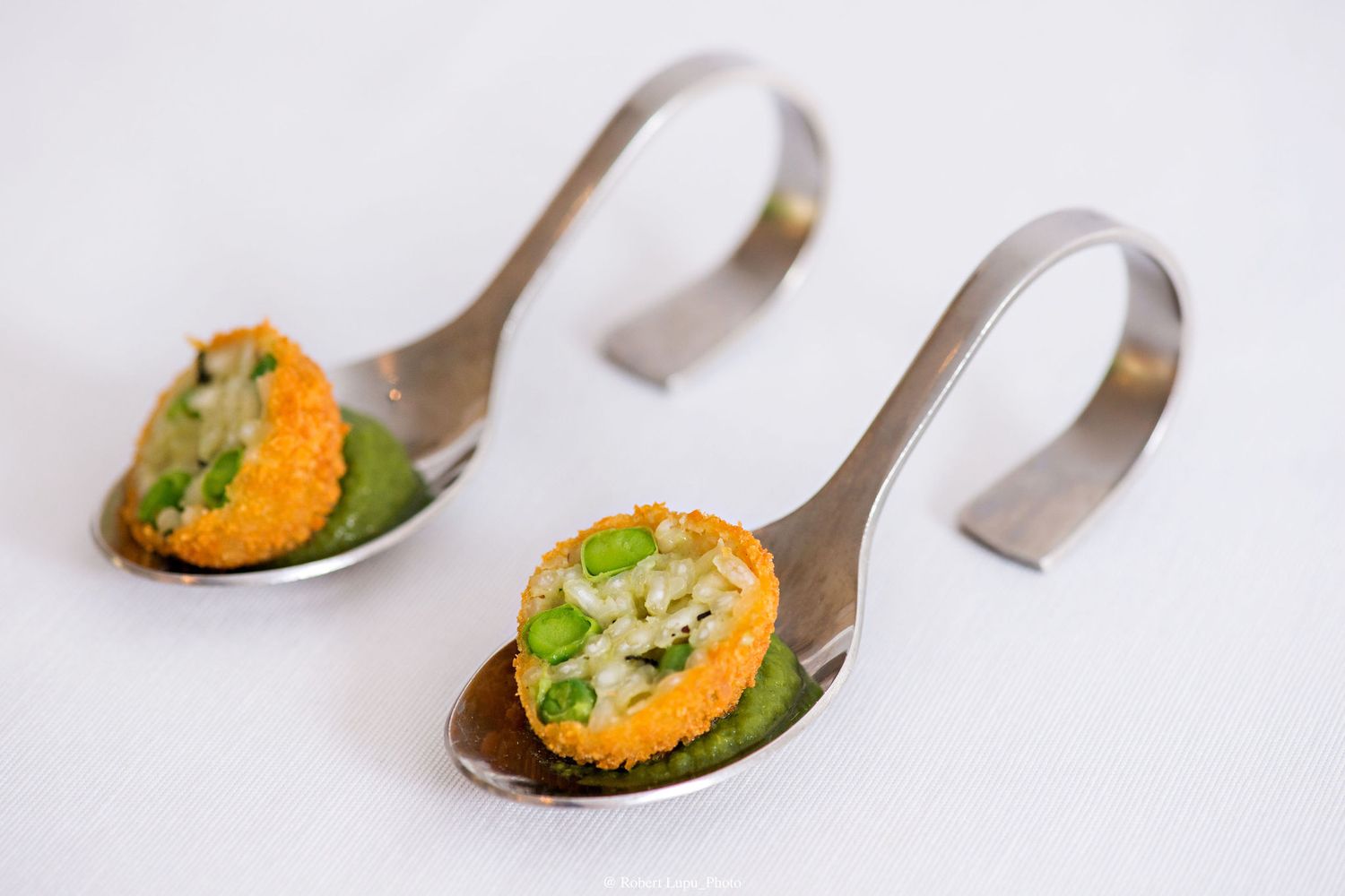 Two elegant silver tasting spoons with green puree topped with crispy orange-coated bites and diced garnish on white background.