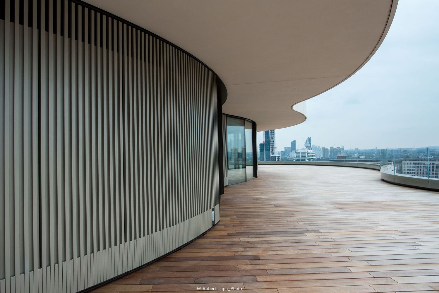 Modern curved building terrace with wooden deck flooring, vertical metal slats, glass railings, and city skyline views on overcast day.