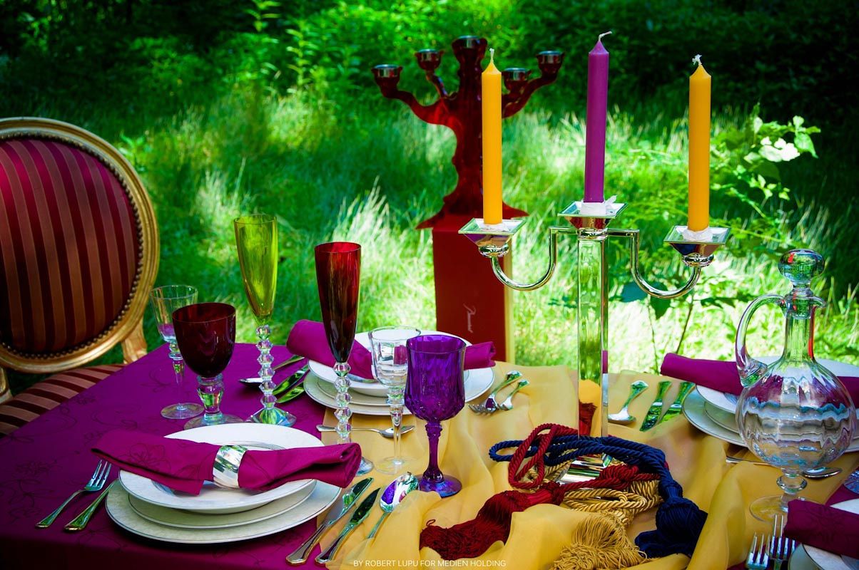 Elegant outdoor dining table with purple linens, colorful candles, fine china, and crystal glasses amid lush green garden backdrop.