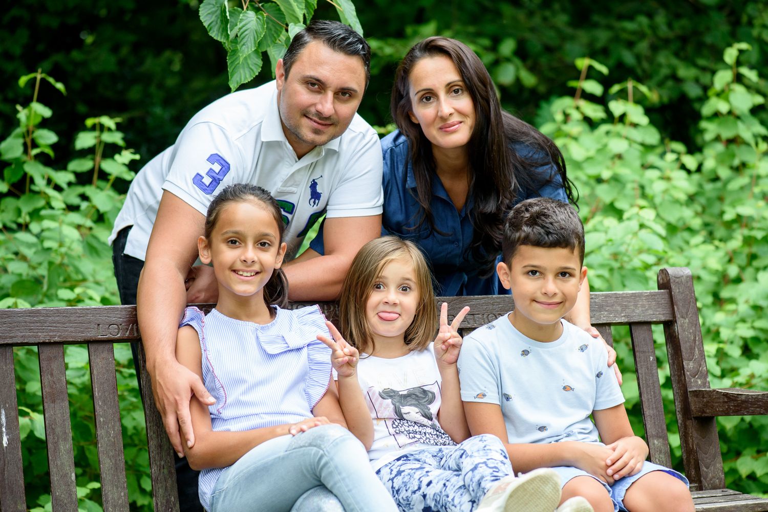 Happy family of five posing on a wooden bench surrounded by lush green foliage in an outdoor park setting.