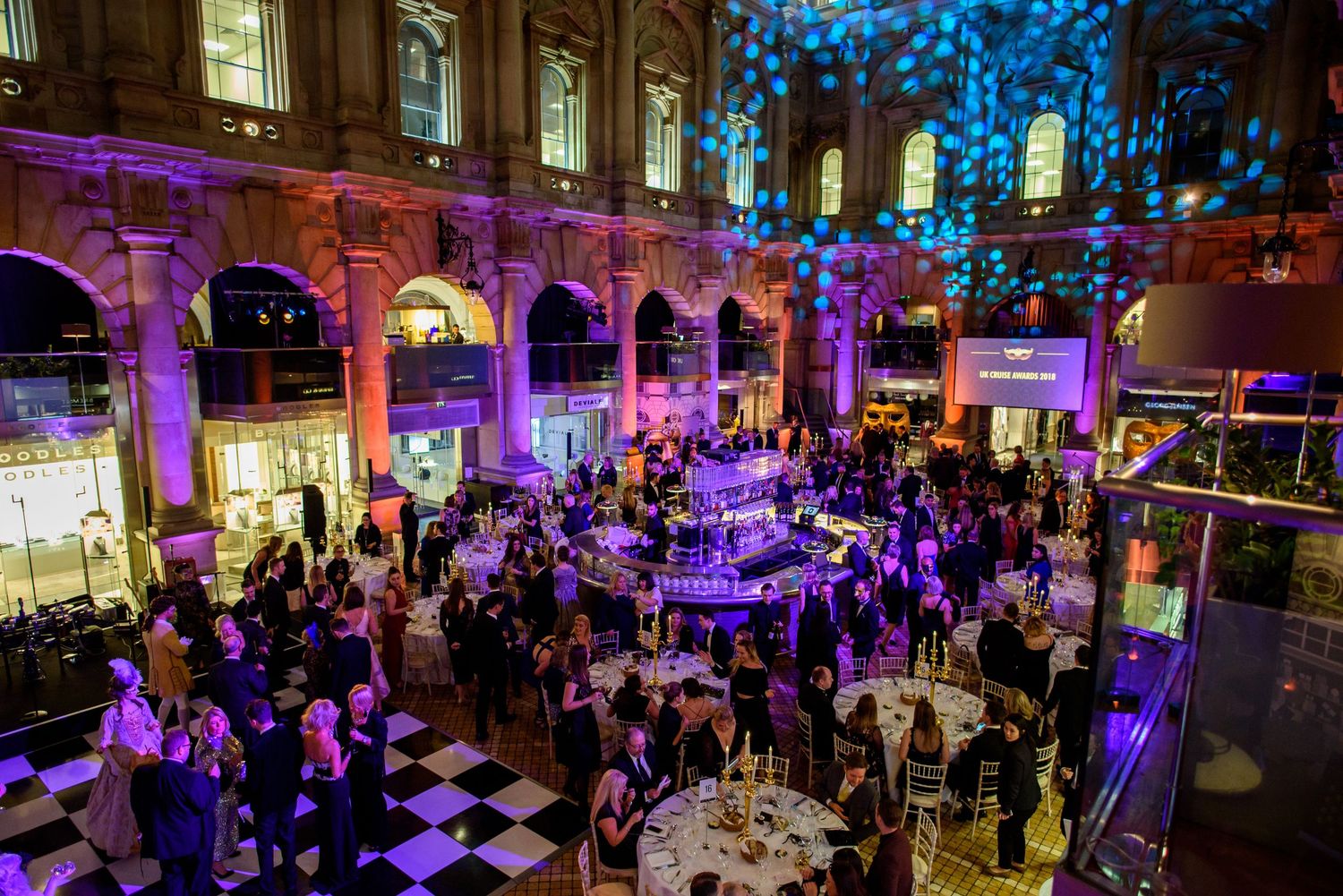 Elegant gala event in a grand historic venue with purple lighting, checkered floors, and guests mingling around illuminated tables.