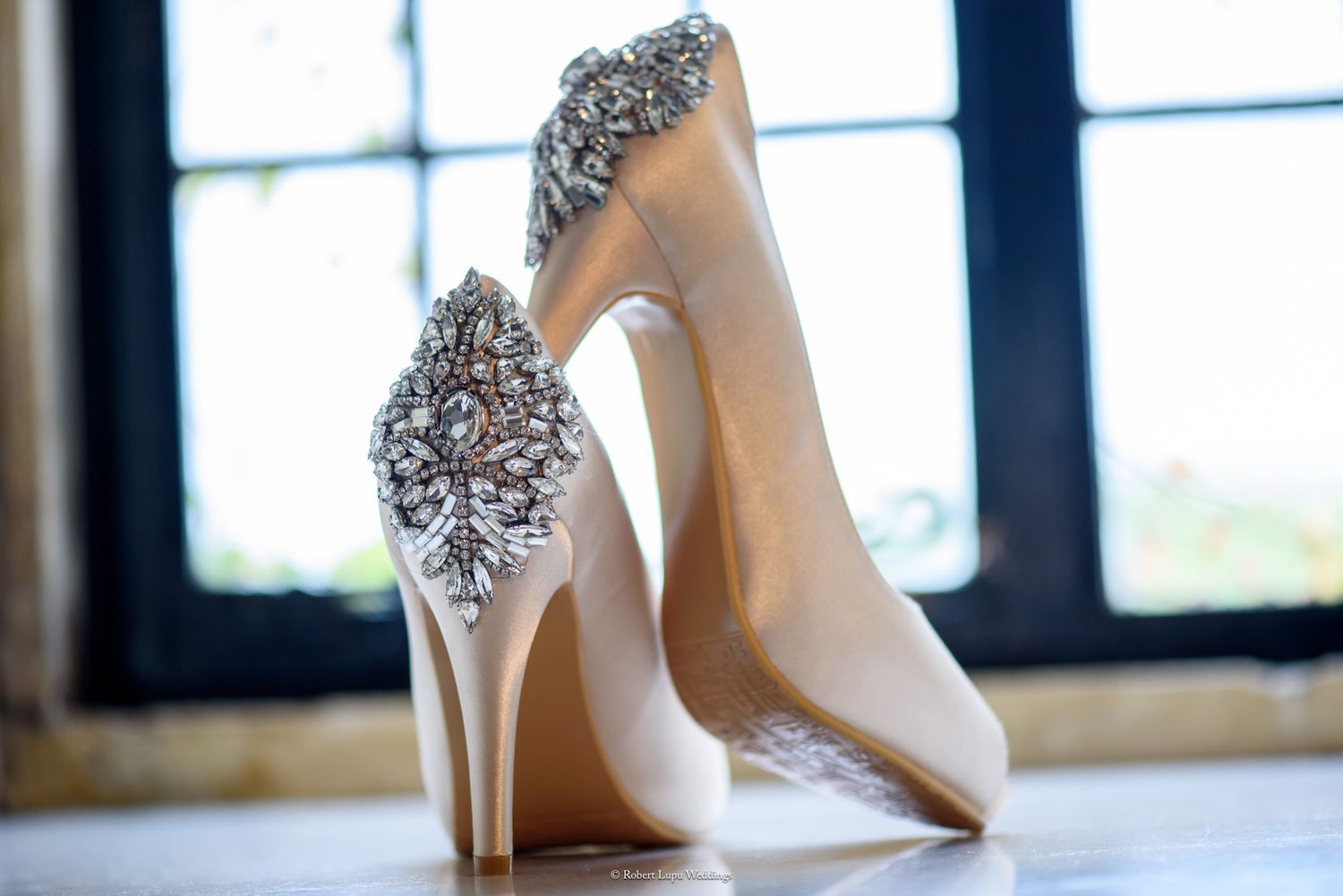 Elegant white bridal heels with sparkling crystal embellishments displayed near a window.