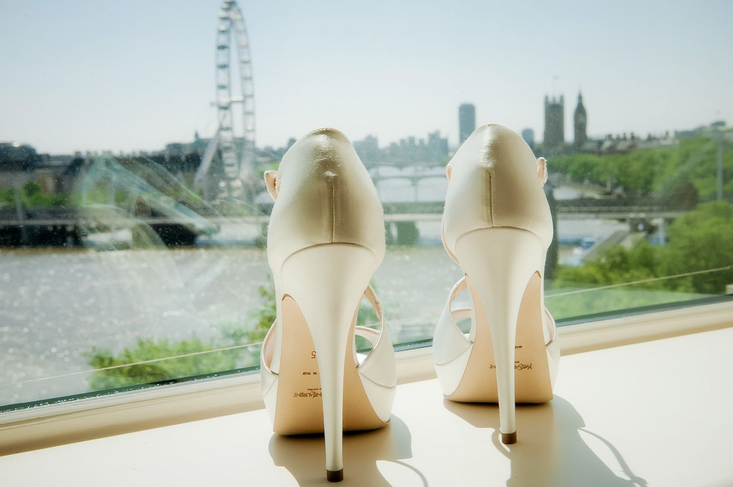 White bridal heels displayed on a windowsill with the London Eye and Westminster skyline in the background.