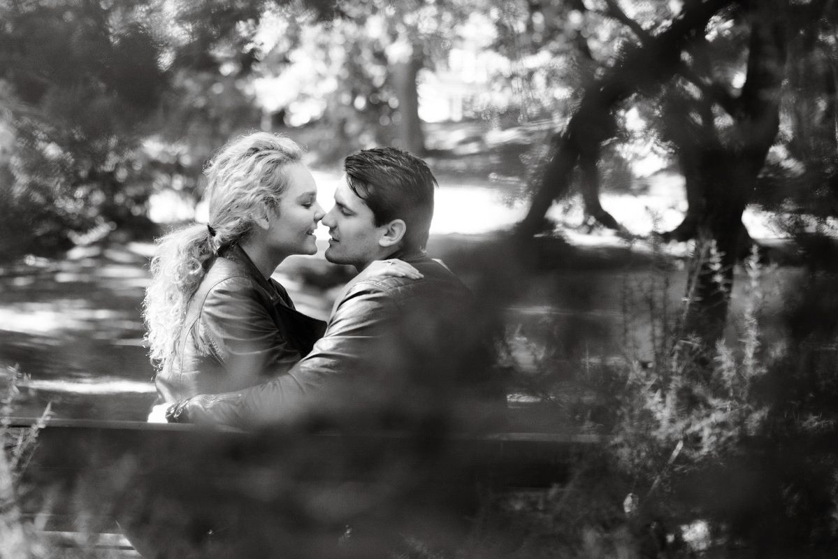 Black and white photo of a romantic couple sharing a tender kiss outdoors under lush tree canopy.