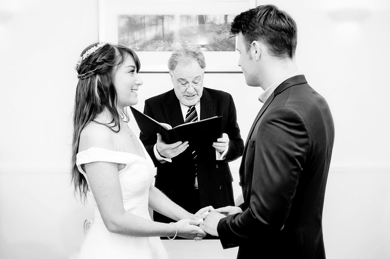 Black and white photo of a couple exchanging vows at a wedding ceremony with an officiant reading from a book.