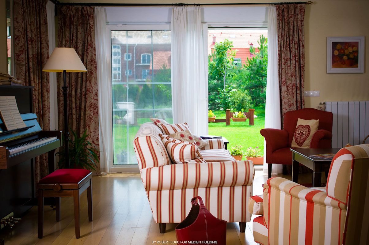Cozy living room with red and white striped sofas, a piano, and large windows overlooking a lush green garden.