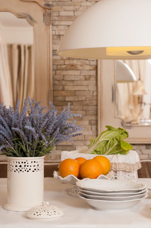 Elegant kitchen counter with lavender bouquet, fresh oranges, romaine lettuce, white plates, and pendant light over brick wall.