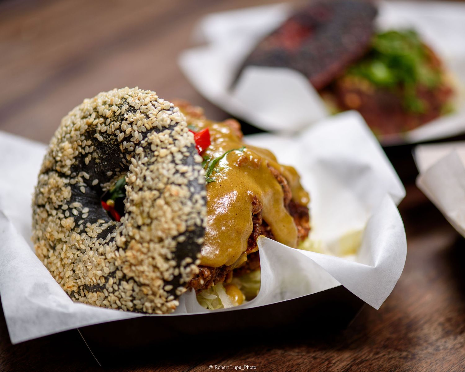 Sesame-crusted burger topped with savory sauce and peppers served in a paper-lined tray at a restaurant.