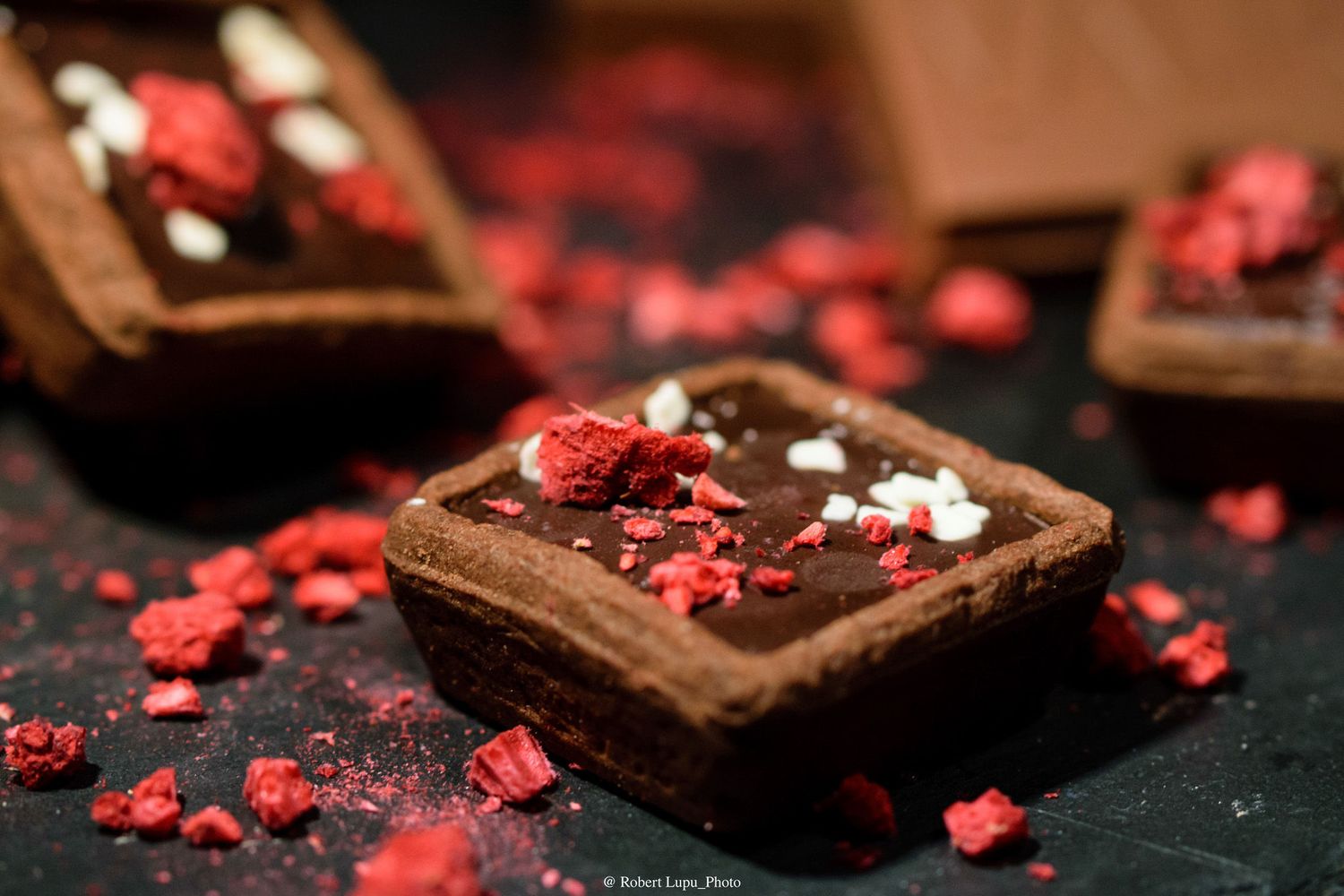 Chocolate brownie tart topped with dark ganache and crushed freeze-dried raspberries on a dark slate surface.