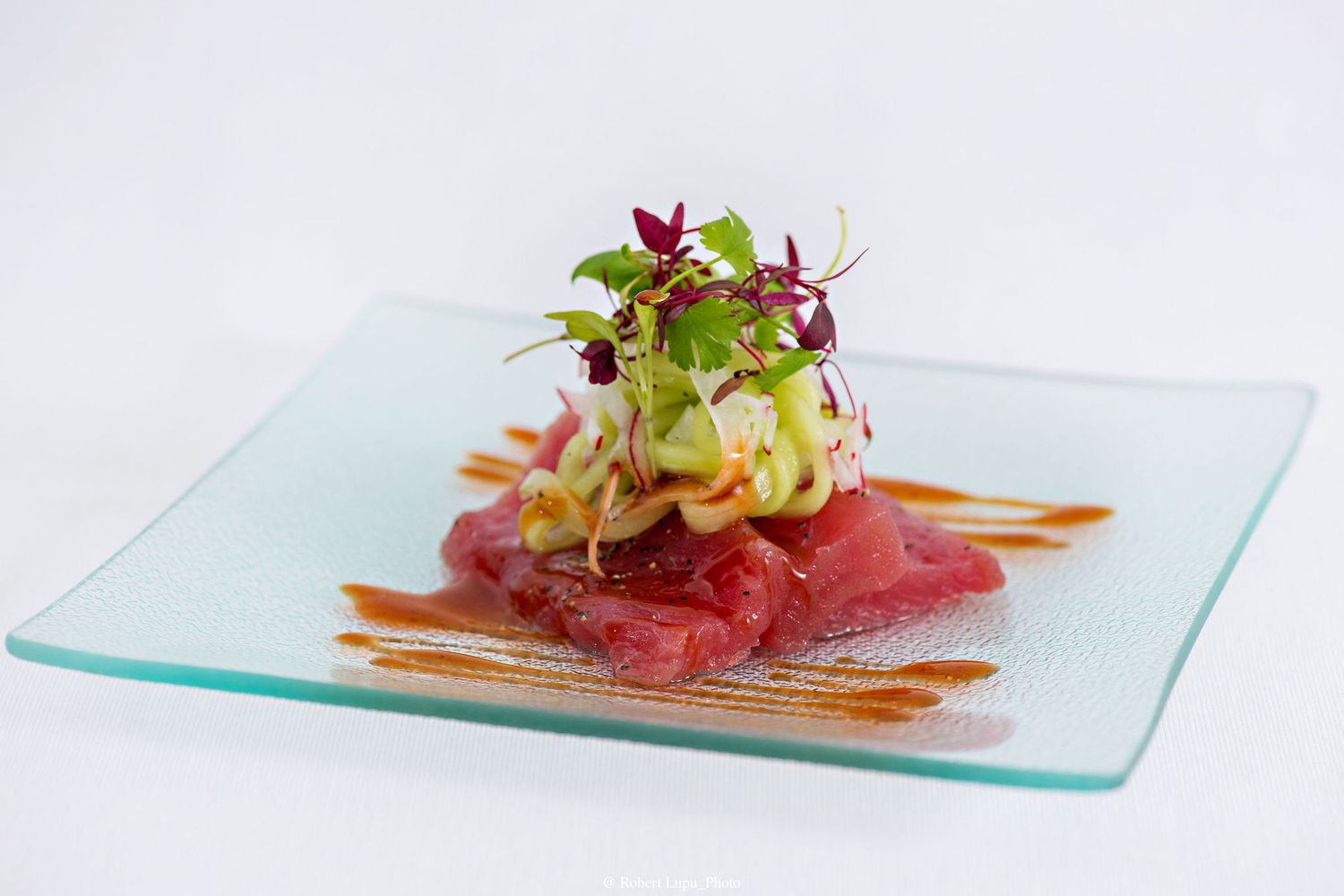 Elegant tuna tartare with avocado and microgreens on a glass plate, drizzled with sauce.