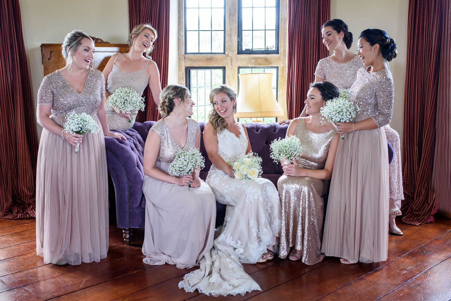 Six bridesmaids in dusty rose gowns holding white bouquets surround a bride in a white lace wedding dress indoors.