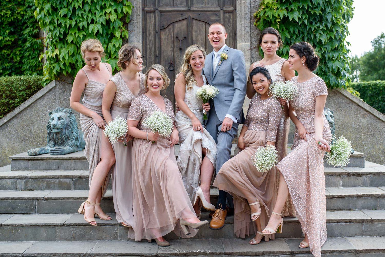 Wedding party poses on stone steps, groom in gray suit with bridesmaids in blush pink gowns holding white floral bouquets.