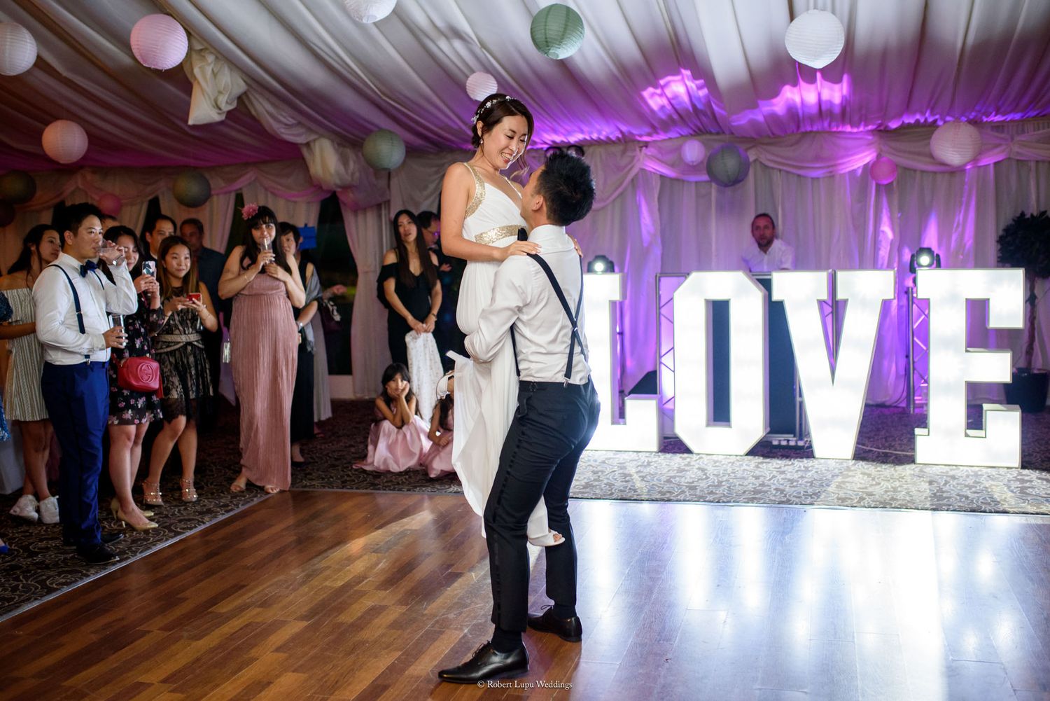 Groom lifts bride during first dance at wedding reception with illuminated LOVE letters in background.