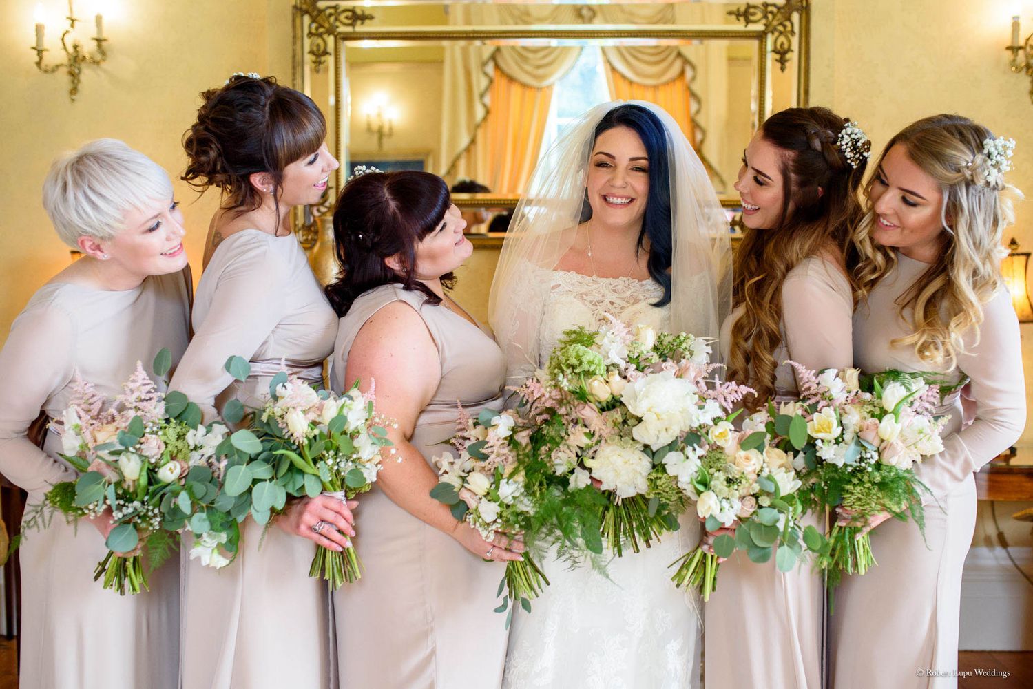 Bride in white gown and veil smiles with five bridesmaids in champagne dresses holding elegant floral bouquets.