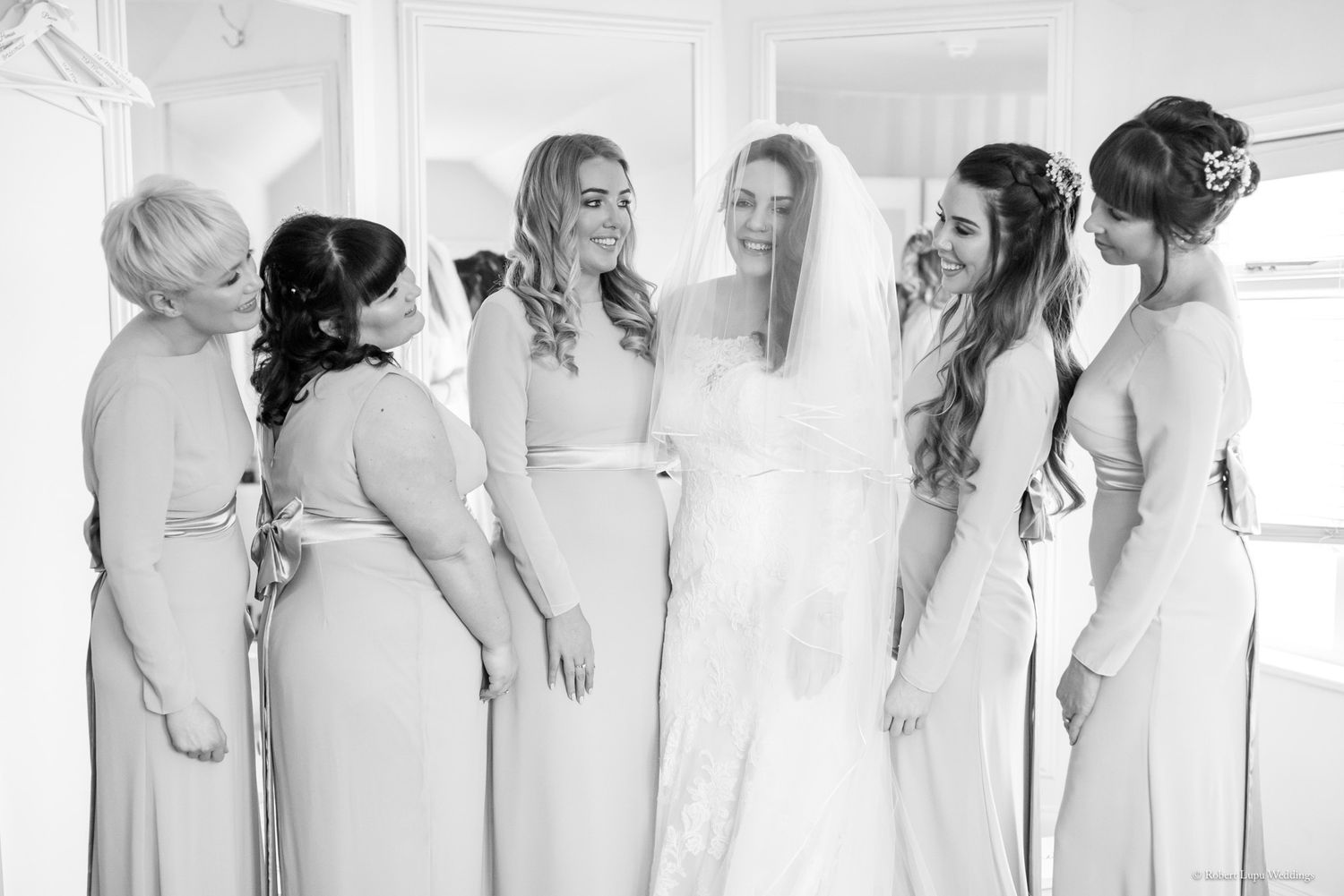 Bride in white gown and veil shares a joyful moment with bridesmaids in champagne dresses.