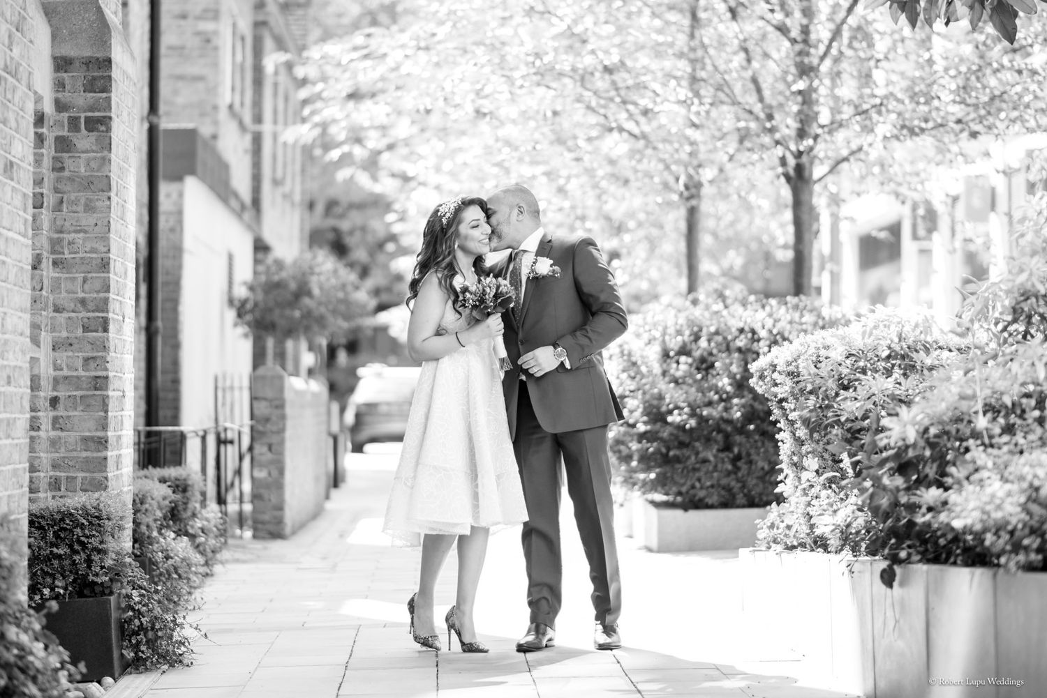 Black and white photo of a couple sharing a romantic kiss on a tree-lined sidewalk in elegant wedding attire.