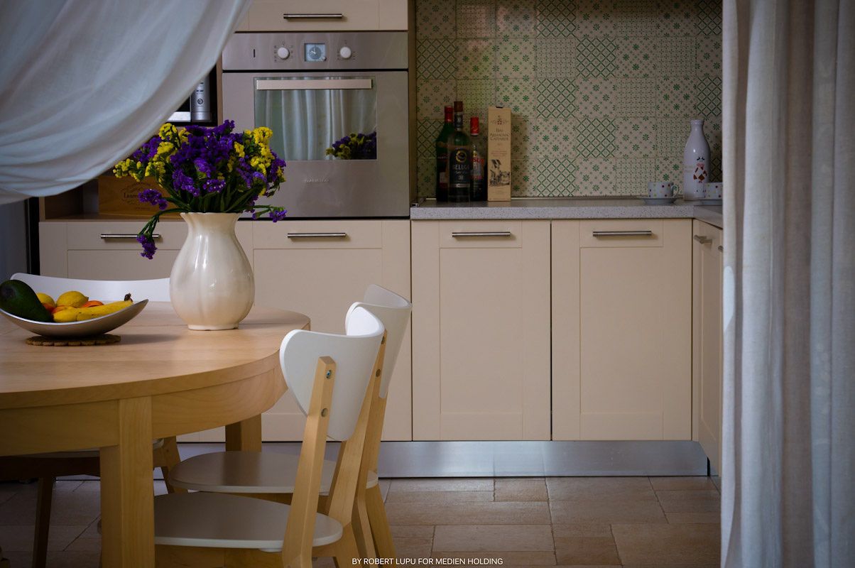 Bright kitchen with mosaic tile backsplash, cream cabinets, and wooden dining table with purple flowers.
