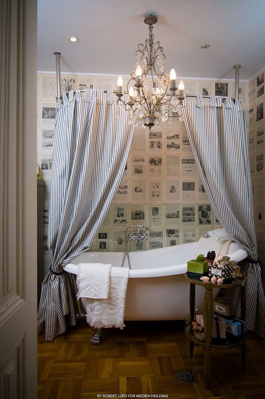 Elegant bathroom with clawfoot tub, striped curtains, chandelier, and newspaper-covered walls on herringbone wood floors.
