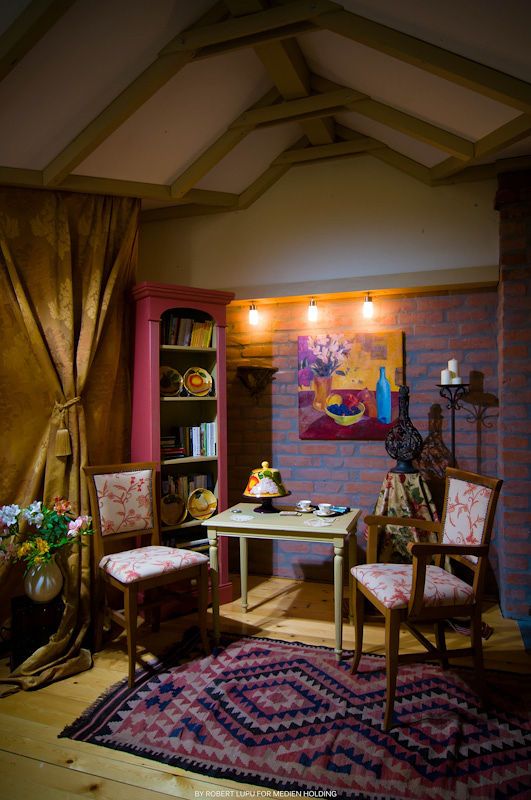 Rustic attic sitting room with brick walls, floral chairs, wooden table, bookshelf, colorful rug, and warm pendant lighting.
