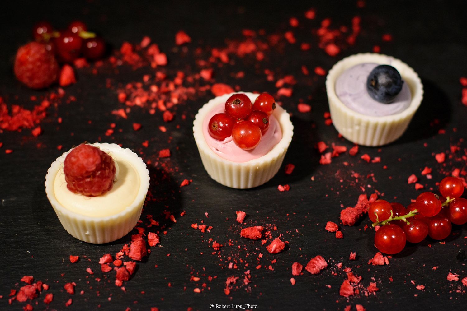 Three elegant mini cheesecakes topped with fresh raspberries, red currants, and blueberries on a dark slate board.